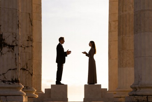 Two people stand on separate stone platforms between classical columns, facing each other in conversation or debate.